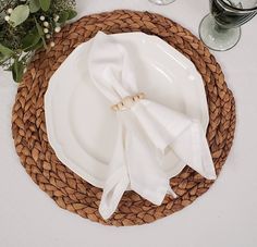 a place setting with white plates and napkins on a woven place mat, surrounded by greenery