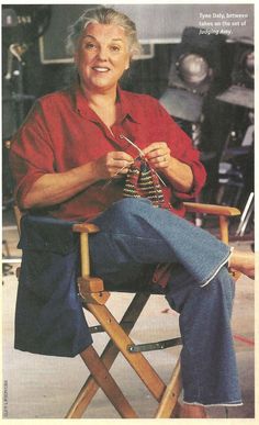 an older woman sitting in a wooden chair with her hands on the knitting needles and smiling