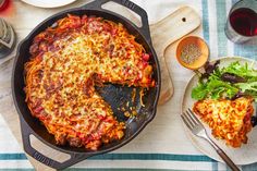 a skillet filled with lasagna next to a plate of salad and wine