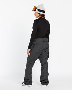 a woman in black shirt and grey pants standing on white background wearing snowboard gear