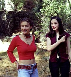 two women standing next to each other in front of trees and bushes, one wearing a red top