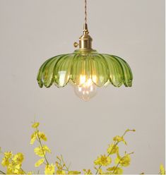 a green light hanging from a ceiling with yellow flowers in the foreground and a white wall behind it