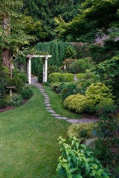 a garden with lots of green grass and trees in the background, along with a stone path that leads to an arbor