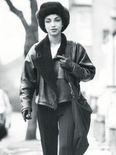 black and white photograph of woman walking down the street wearing a fur hat, leather jacket, and pants