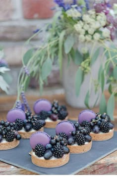 small desserts with blackberries and blueberries are arranged on a slate tray next to flowers