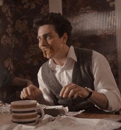 two men sitting at a table eating cake and drinking tea, one man is holding a fork in his mouth