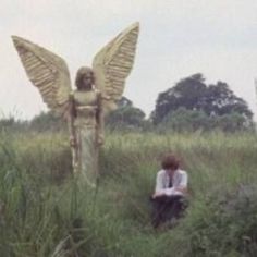 an angel statue sitting in the middle of a field next to a woman with wings