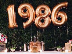 a table topped with lots of gold balloons and flowers next to a wall covered in greenery