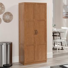 a tall wooden cabinet next to a trash can in a room with white walls and wood flooring