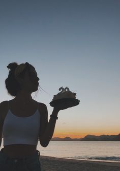 a woman standing on the beach with a cake in her hand and sunset behind her