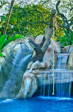 an artificial waterfall in the middle of a pool surrounded by trees and rocks with water running down it