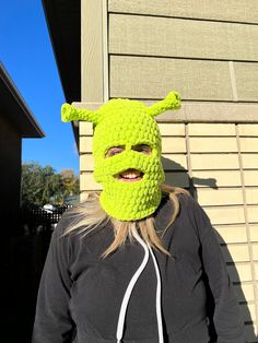 a woman wearing a green crochet mask and black hoodie standing in front of a house