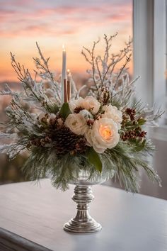 a vase filled with white flowers and pine cones on top of a table next to a window