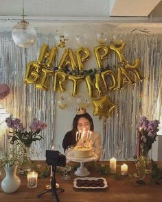 a woman sitting in front of a birthday cake with candles on it and balloons hanging from the ceiling