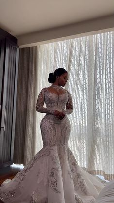 a woman standing in front of a window wearing a wedding dress with long sleeves and beading