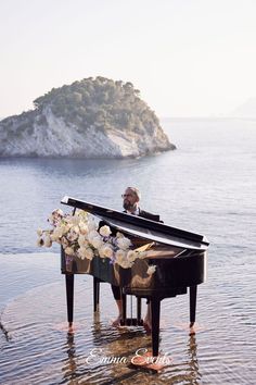 a man sitting at a piano in the water with flowers on his feet and an island in the background