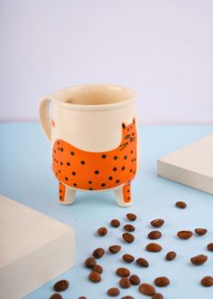 a ceramic cat mug with coffee beans scattered around it on a blue surface next to a white box