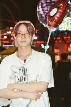 a young man with glasses and a spiderman balloon on his head stands in front of an amusement park at night