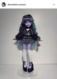 a doll with blue hair and purple shoes is standing on a white surface in front of a wall