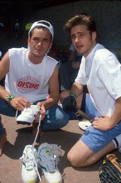 two men sitting next to each other on the ground with shoes around them and one man tying his shoelaces