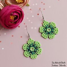 two green and blue beaded earrings sitting next to a pink flower on a table