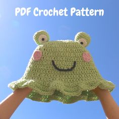 a crocheted frog hat is held in the air by someone's hands