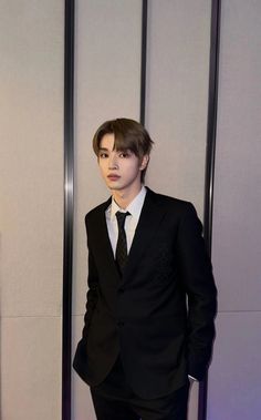 a young man in a suit and tie standing next to a tall metal wall with his hands in his pockets