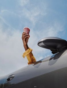 a person holding up a coffee cup in front of a car window with blue sky and clouds behind them