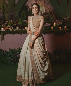 a woman in a white and gold lehenga standing on the grass with flowers behind her