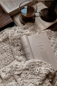 an open book sitting on top of a blanket next to a cup of coffee