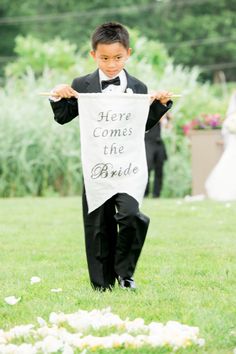 a young boy in a tuxedo holds up a sign that says here comes the bride