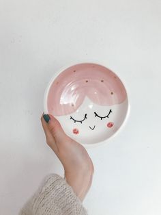 a hand holding a pink and white bowl with a face painted on the side, in front of a white wall