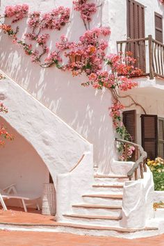 a white building with pink flowers growing on it's side and stairs leading up to the upper floor