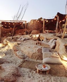 an outdoor seating area is lit up with string lights and pillows on the sand at dusk