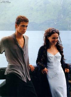 a man and woman standing next to each other on a pier near the water with mountains in the background