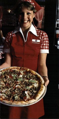 a woman in an apron holding a pizza on a platter with toppings and smiling