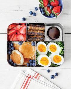 a bento box filled with eggs, fruit and sausages next to a bowl of strawberries