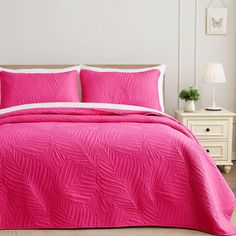 a bed with pink comforter and pillows in a white room next to a night stand