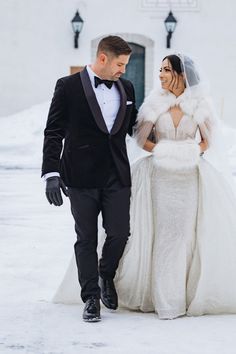 a bride and groom walking together in the snow at their winter wedding ceremony, dressed in black tuxedos