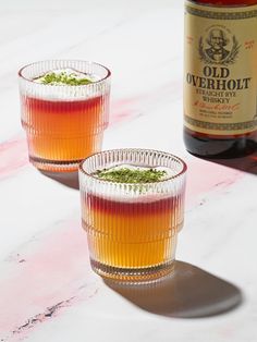 two shot glasses filled with liquid next to a bottle of alcohol on a marble surface