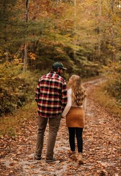 a man and woman walking down a leaf covered path in the woods with trees behind them