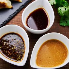 three white bowls filled with sauce on top of a wooden table next to bread and cilantro