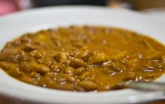 a white bowl filled with beans and meat on top of a wooden table next to a fork