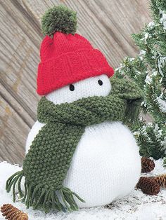 a knitted snowman wearing a red hat and scarf with pine cones around it