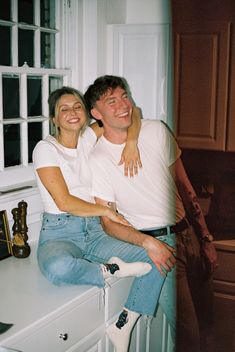 a man and woman sitting on top of a counter in a kitchen smiling at the camera