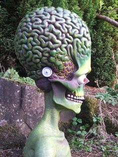 a green statue with a brain on it's head in the middle of some bushes