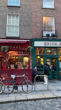 two bicycles are parked in front of a brick building on a street corner with people sitting outside