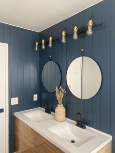 a bathroom with two sinks, mirrors and lights on the wall above it is blue painted wood paneling