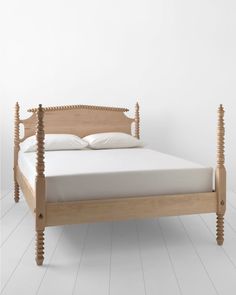 a wooden bed frame with white sheets and pillows on it, in front of a white wall