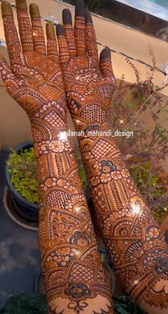 two hands that are decorated with henna and some plants in the background, one is holding